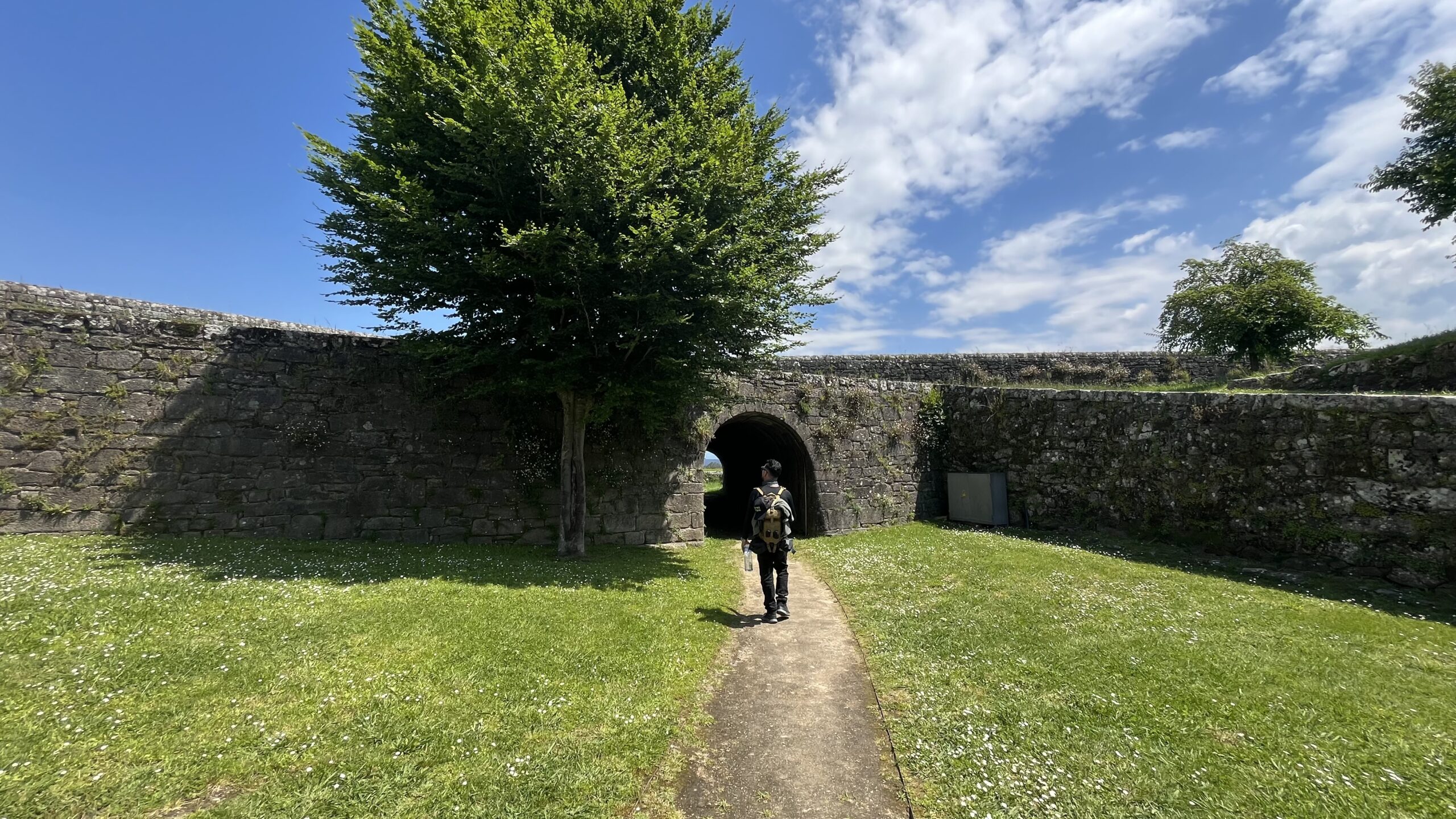Caminhante de mochila atravessando um arco de pedra num muro histórico, no Caminho Português.